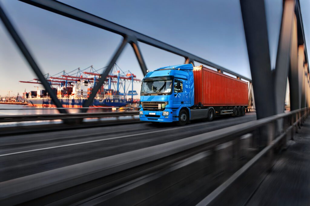 Containertransport met vrachtwagen op de brug bij de haven in Rotterdam, symboliseert logistiek, supply chain en internationaal verkeer, met havenkranen op de achtergrond.