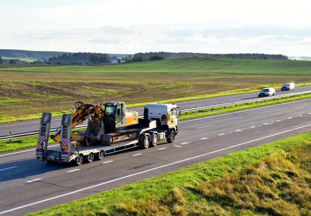 Beton heftruck op vrachtwagen die op snelweg geladen en vervoerd wordt, met groene velden en uitgestrekte landschap op de achtergrond.