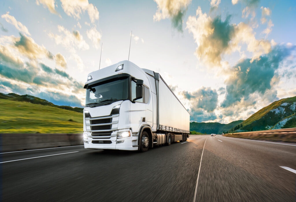 Snelle witte vrachtwagen rijdend over snelweg met groene heuvels en blauwe lucht in de achtergrond.