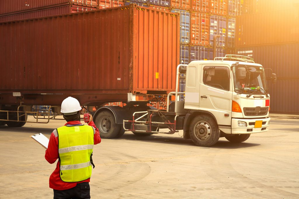 Veiligheidsspecialist controleert containerlogistiek op transportterrein met vrachtwagen en gestapelde containers.