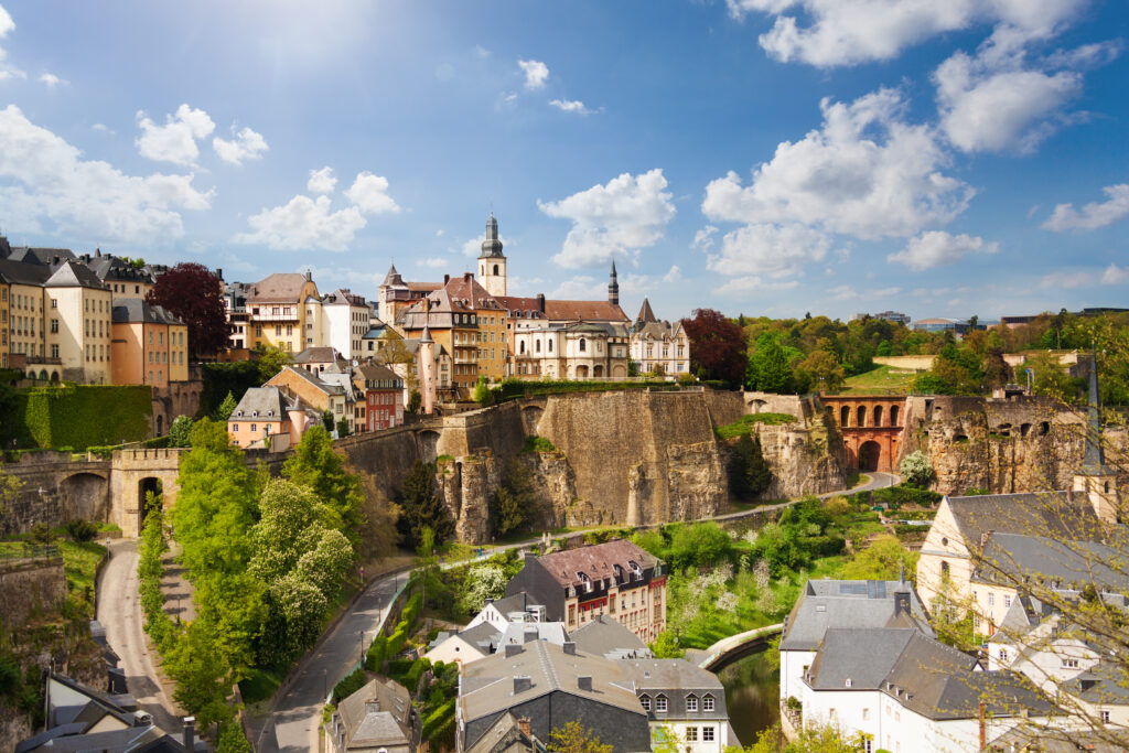 Gezicht op een historisch stadje met kasteelmuur, kleurrijke gebouwen, kerktorens en groene heuvels in Luxemburg, onder een heldere blauwe lucht met wolken.