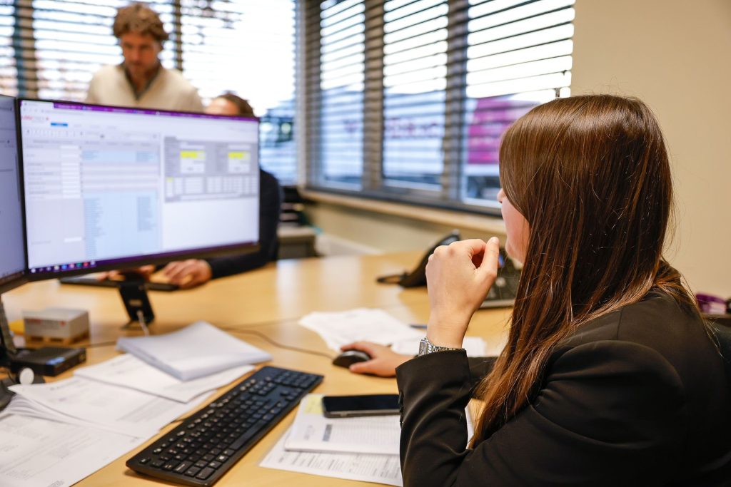 Efficiënt kantoorwerk met vrouw die digitaal documenten beheert op computer, te midden van papieren en kantoorbenodigdheden, in een moderne werkruimte met grote ramen en natuurlijke lichtinval.